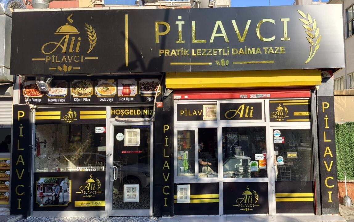 Ali Pilavcı'nın Kartal Karlıktepe'deki dükkânı önden görünüyor: siyah zemin üzerine altın harflerle Ali PİLAVCI tabelası, altında sarı şerit, vitrinde yemek fotoğrafları ve cam giriş kapısı